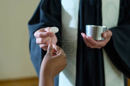 Foto de Mano de una joven feligresa que toma pan sin levadura sostenido por un sacerdote católico en sotana durante la comunión después de la liturgia - Imagen libre de derechos