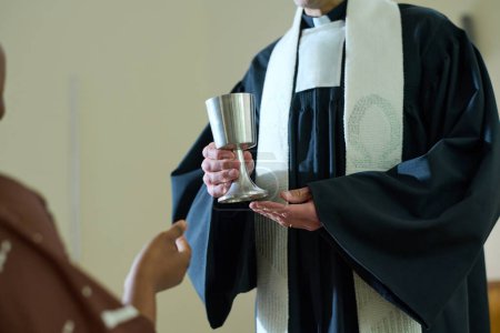 Photo for Clergyman of Catholic church holding cup with wine for oblation rite while standing in front of African American female parishioner - Royalty Free Image