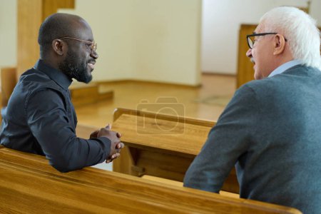 Photo for Young African American pastor consulting senior male parishioner after church service while both sitting on bench in front of one another - Royalty Free Image