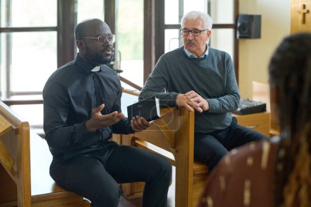 Téléchargez les photos : Pasteur confiant en chemise noire avec col clérical expliquant les versets de la Bible aux paroissiens interculturels de son église après le sermon - en image libre de droit