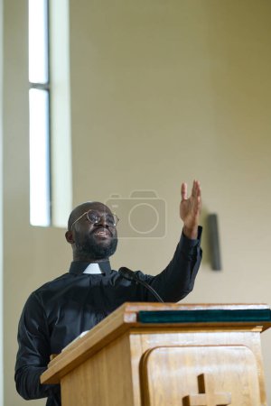 Téléchargez les photos : Jeune prêtre en chemise noire avec col clérical debout à la chaire et parlant au microphone tout en gardant le bras levé en geste de bénédiction - en image libre de droit