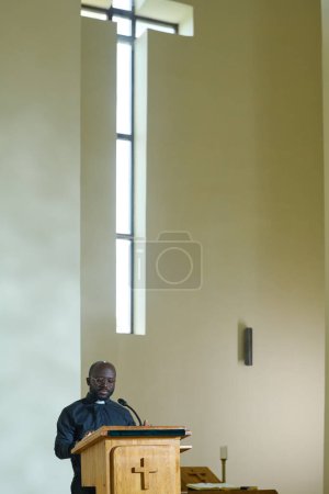 Photo for Young black man standing by wooden pulpit in front of parishioners while reading Bible aloud and commenting verses during church service - Royalty Free Image