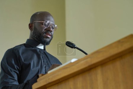 Photo for Young priest in black shirt with clerical collar reading verses from Bible and explaining them while pronouncing speech by pulpit - Royalty Free Image