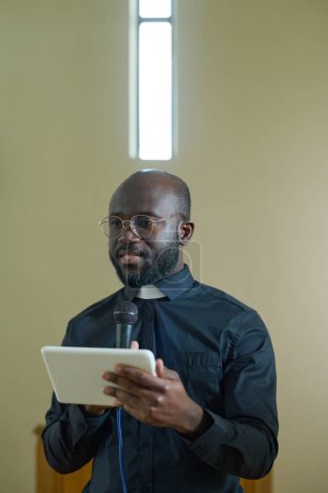 Foto de Pastor con micrófono y tableta usando la Biblia electrónica o en línea mientras pronuncia sermón frente a los feligreses durante el servicio a la iglesia - Imagen libre de derechos