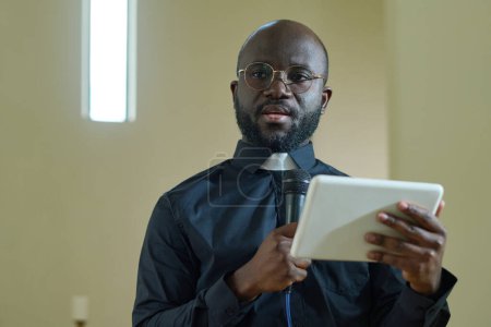 Téléchargez les photos : Jeune homme noir confiant avec microphone et tablette numérique faisant la parole pour les paroissiens de l'église évangélique pendant le sermon - en image libre de droit