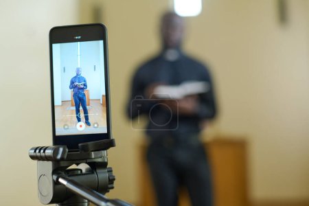 Smartphone con pastor afroamericano en pantalones negros y camisa con cuello clerical sosteniendo la Biblia abierta en pantalla durante la transmisión del sermón