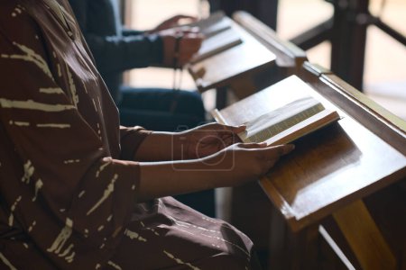 Téléchargez les photos : Jeune femme noire assise près d'un bureau en bois dans une église catholique ou évangélique et étudiant la Sainte Bible au séminaire - en image libre de droit