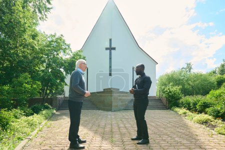 Photo for Side view of young pastor and senior male parishioner having discussion in church yard while standing in front of one another - Royalty Free Image