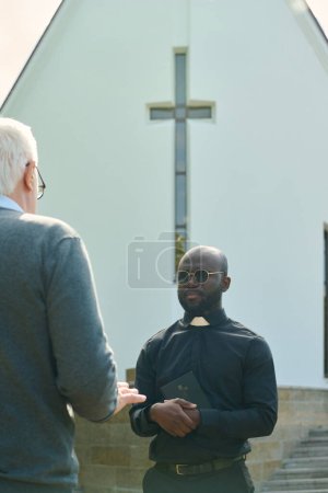 Photo for Young serious priest in black shirt with clerical collar holding Holy Bible by chest and talking to aged male parishioner against church building - Royalty Free Image