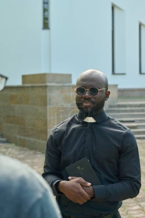 Téléchargez les photos : Jeune pasteur afro-américain en chemise noire avec col clérical consultant paroissien tout en se tenant dans la cour de l'église contre le bâtiment blanc - en image libre de droit