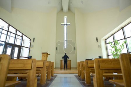 Foto de Interior de la iglesia con pasillo largo que conduce al pastor de pie delante de la cruz y el púlpito de madera y mantener sus brazos en gesto de bendición - Imagen libre de derechos