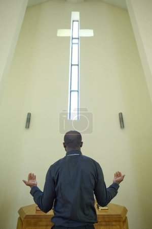 Foto de Vista trasera del joven pastor afroamericano vestido de negro rezando mientras está de pie junto al altar o el púlpito frente a la cruz - Imagen libre de derechos
