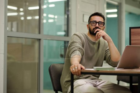 Téléchargez les photos : Jeune homme d'affaires coûteux avec des lunettes assis par bureau devant un ordinateur portable et brainstorming tout en travaillant sur un nouveau projet - en image libre de droit