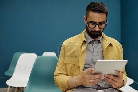 Photo for Young serious male employee with beard looking through online information on screen of tablet while preparing report - Royalty Free Image