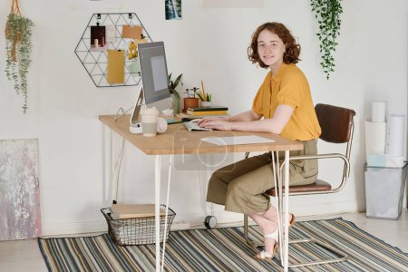 Photo for Happy young female freelancer in casualwear sitting by desk in front of computer monitor in living room and looking at camera - Royalty Free Image
