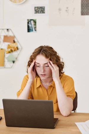 Téléchargez les photos : Jeune femme d'affaires exprimant malentendu ou fatigue tout en étant assis par bureau devant un ordinateur portable et en regardant à travers les données en ligne - en image libre de droit