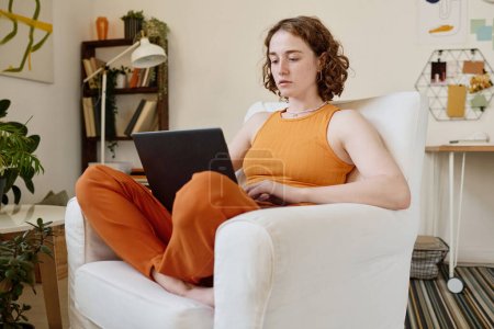 Foto de Joven empresaria seria con portátil en sus rodillas concentrándose en la red mientras está sentada en un cómodo sillón de cuero blanco - Imagen libre de derechos
