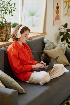 Photo for Youthful female student in red shirt and white pants typing on laptop keyboard while sitting on soft comfortable couch in living room - Royalty Free Image