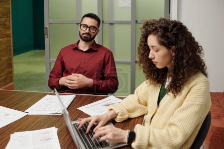 Téléchargez les photos : Confidenr homme d'affaires assis devant une collègue féminine tapant sur le clavier de l'ordinateur portable tout en travaillant sur un nouveau projet d'entreprise - en image libre de droit