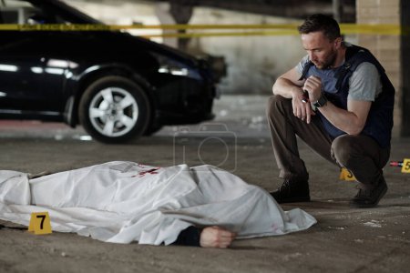 Foto de Joven detective masculino con linterna pequeña inspeccionando el área de la escena del crimen con un cadáver cubierto rodeado de tarjetas amarillas con números - Imagen libre de derechos