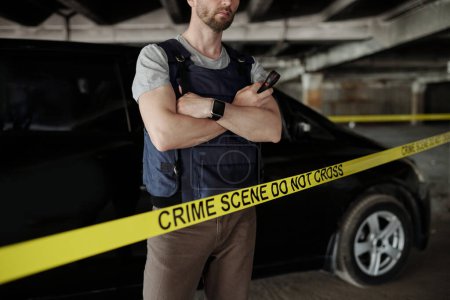 Photo for Cropped shot of crime scene investigator in bulletproof vest crossing arms by chest while standing behind yellow tape against car - Royalty Free Image