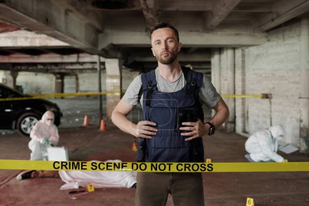 Photo for Young serious policeman in bulletproof vest standing behind crime scene tape and looking at camera against forsenic experts working - Royalty Free Image