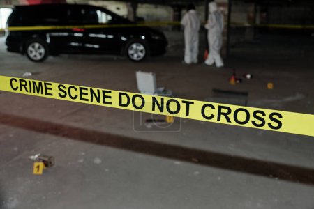 Photo for Yellow tape surrounding crime scene in parking area with two experts in protective coveralls seeking for evidences of murder - Royalty Free Image