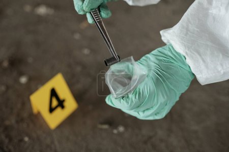 Foto de Guante manos de experto criminológico con pinzas metálicas poniendo cartucho vacío en el paquete de plástico durante la inspección de evidencias - Imagen libre de derechos