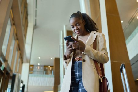 Foto de Joven mujer de negocios afroamericana elegante con teléfono móvil hablando a través de chat de vídeo mientras está de pie en el salón del hotel - Imagen libre de derechos