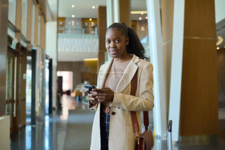 Foto de Mujer afroamericana joven en ropa de casualwear inteligente mirando a la cámara mientras está de pie en el salón del hotel y mensajes de texto en el teléfono móvil - Imagen libre de derechos
