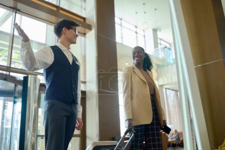 Photo for Owner of receptionist of hotel in uniform meeting black woman with suitcase and telling her where she can find receptionist - Royalty Free Image