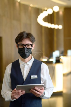 Téléchargez les photos : Jeune homme d'affaires confiant ou travailleur de l'hôtel moderne en uniforme, lunettes et masque protecteur debout devant la caméra - en image libre de droit