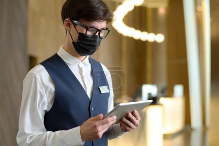 Photo for Young receptionist of modern hotel in uniform, eyeglasses and protective mask using tablet while looking through online reservations - Royalty Free Image