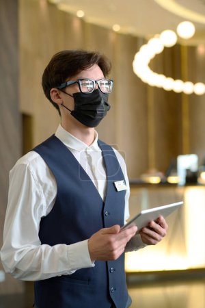 Téléchargez les photos : Jeune réceptionniste sérieux avec tablette debout dans le salon spacieux de l'hôtel moderne de luxe tout en rencontrant de nouveaux clients - en image libre de droit