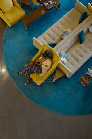 Foto de Por encima de tiro de joven mujer negra con gadget móvil relajante en sillón por dos sofás con cojines en el salón del hotel - Imagen libre de derechos