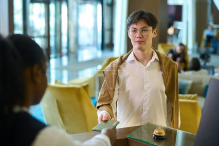 Téléchargez les photos : Jeune homme d'affaires sérieux d'origine asiatique passant son passeport à la réceptionniste au comptoir lors de l'enregistrement dans un hôtel de luxe - en image libre de droit