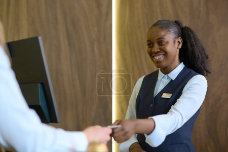 Photo for Happy young black woman in uniform passing passport or keycard to male traveler over counter after registration of his personal data - Royalty Free Image