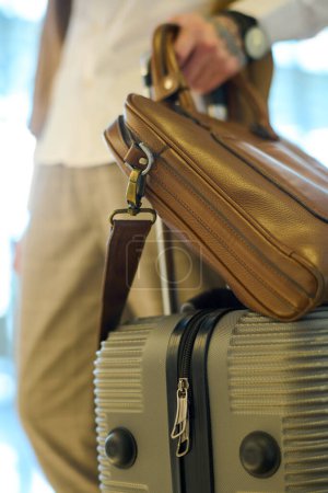 Téléchargez les photos : Valise avec bagages et sac à main en cuir marron tenue par un jeune voyageur debout dans le salon de l'hôtel ou dans le hall de l'aéroport - en image libre de droit