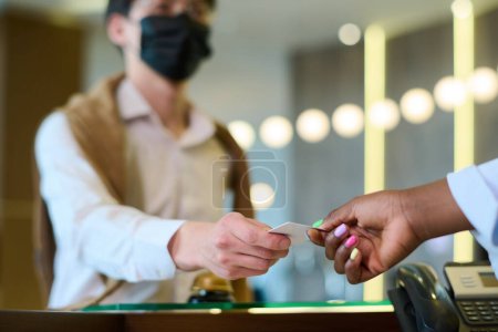 Téléchargez les photos : Main de jeune femme noire prenant la carte-clé tenue par l'invité masculin en casual et masque de protection sur le comptoir d'accueil - en image libre de droit