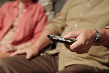 Téléchargez les photos : Main de l'homme âgé en casual-wear en appuyant sur les boutons de télécommande tout en étant assis à côté de sa femme et en choisissant la chaîne de télévision à lesiure - en image libre de droit