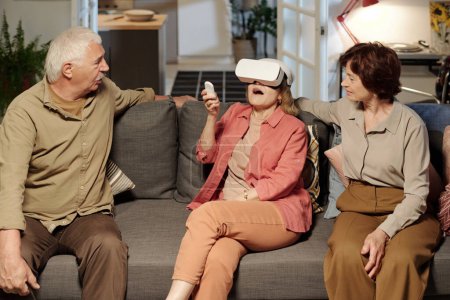 Téléchargez les photos : Heureuse femme âgée dans le casque vr appuyant sur le bouton sur la télécommande pendant le jeu virtuel ou la présentation parmi ses amis - en image libre de droit