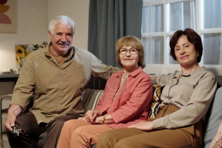 Photo for Group of senior cheerful man and two women in casual shirts and pants relaxing on couch in living room and looking at camera - Royalty Free Image