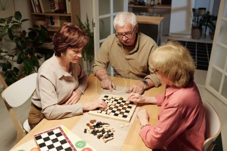 Photo for Three retired friends in casualwear gathered by table to play chess or checkeres, senior woman thinking of next move - Royalty Free Image