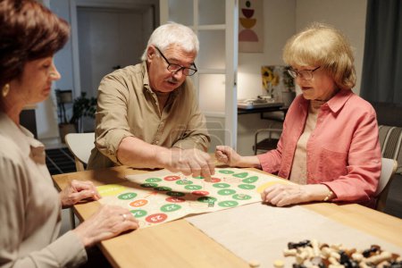 Photo for Senior man moving chip throughout paper board with green and red circles while playing leisure game with two mature women - Royalty Free Image