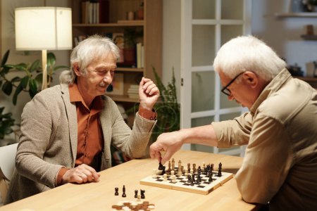 Photo for Happy senior man looking at his buddy moving black figure along chessboard while both playing chess by table at leisure - Royalty Free Image