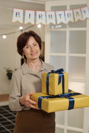 Photo for Aged cheerful woman with packed birthday gifts standing by door of living room and looking at camera while enjoying home party - Royalty Free Image