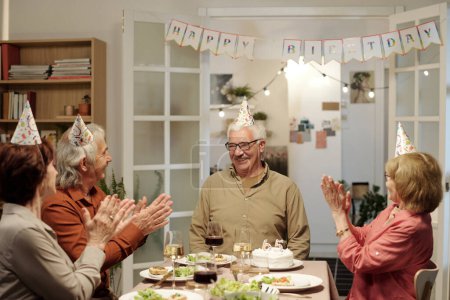 Foto de Feliz hombre mayor mirando a sus amigos dando palmadas en la mesa servida con comida casera durante la celebración del cumpleaños - Imagen libre de derechos