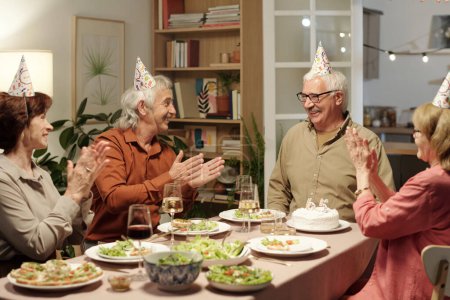 Photo for Group of cheerful senior people clapping hands while looking at their friend celebrating birthday while sitting by festive table - Royalty Free Image
