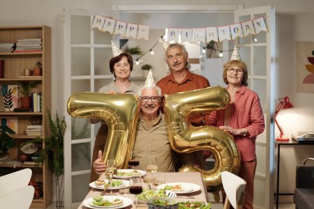 Téléchargez les photos : Groupe d'amis seniors tenant des nombres gonflables montrant l'âge de l'un d'eux tout en se tenant devant la table de fête servie avec de la nourriture maison - en image libre de droit