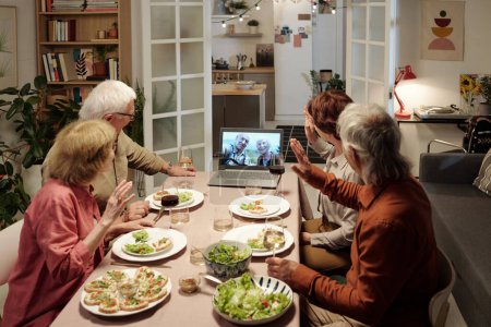 Photo for Group of senior friends waving hands while greeting aged couple on screen of laptop during communication in video chat by table - Royalty Free Image